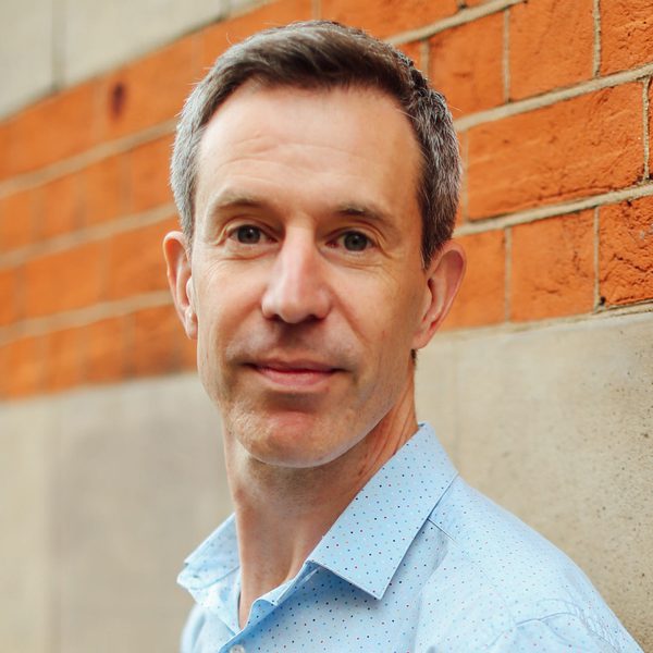Portrait of Julian West looking to camera, smiling, against a brick wall.