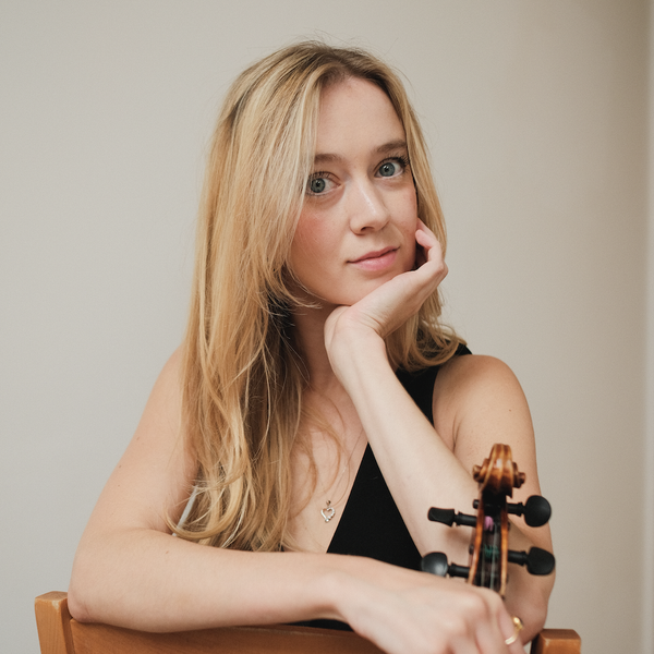 A woman sits backwards, leaning over the backrest of a chair, holding a violin and looking to camera.
