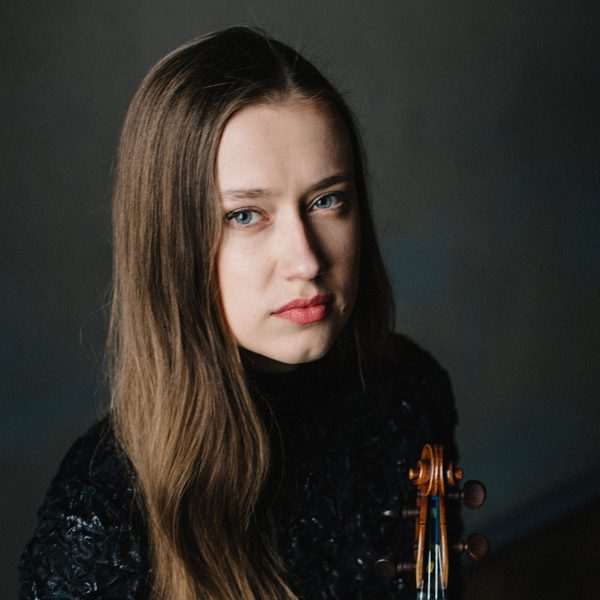 Kristīne Balanas looks to camera, holding violin.