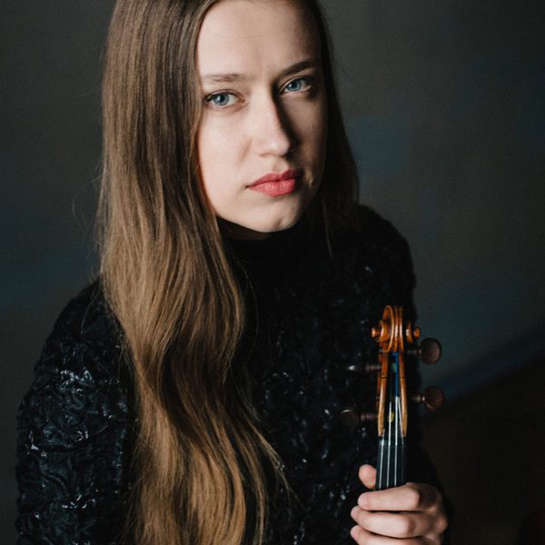 Kristīne Balanas looks to camera, holding violin.