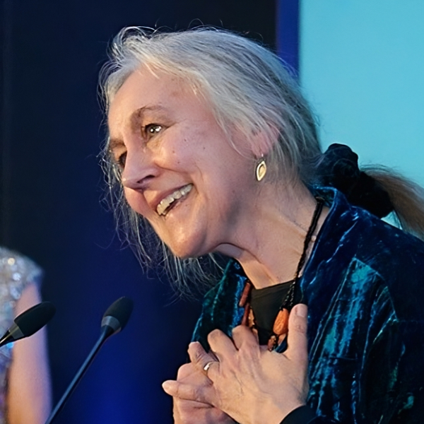 Krystyna Budzynska Stands at a Microphone against a blue background.