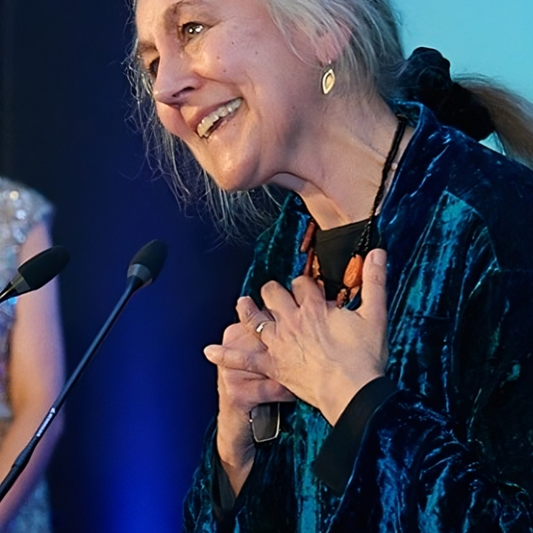 Krystyna Budzynska Stands at a Microphone against a blue background.