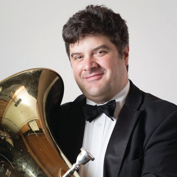 Lee Tsarmaklis holds a tuba, wearing tuxedo, smiling to camera.