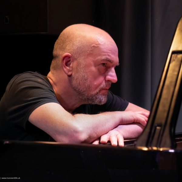 Portrait of Liam Noble sat leaning across the top of a piano, looking off camera.