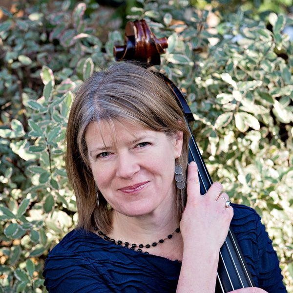 A double bassist supports the neck of her instrument and smiles to camera backed by sunny greenery