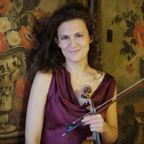Lorenza Borrani smiles holding a a violin, standing in front of a tapestry.