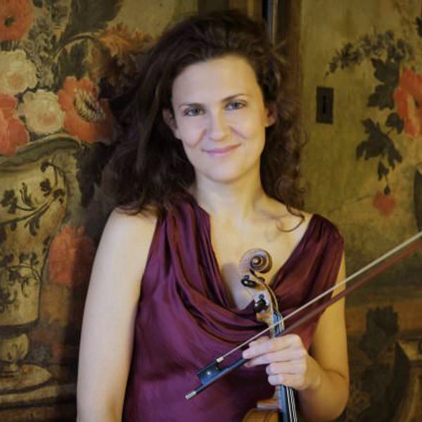Lorenza Borrani smiles holding a a violin, standing in front of a tapestry.