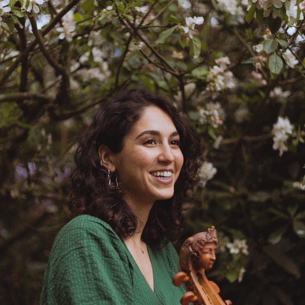 A woman in a green dress, pictured smiling, with the neck of a stringed instrument in the foreground and a tree in the background.
