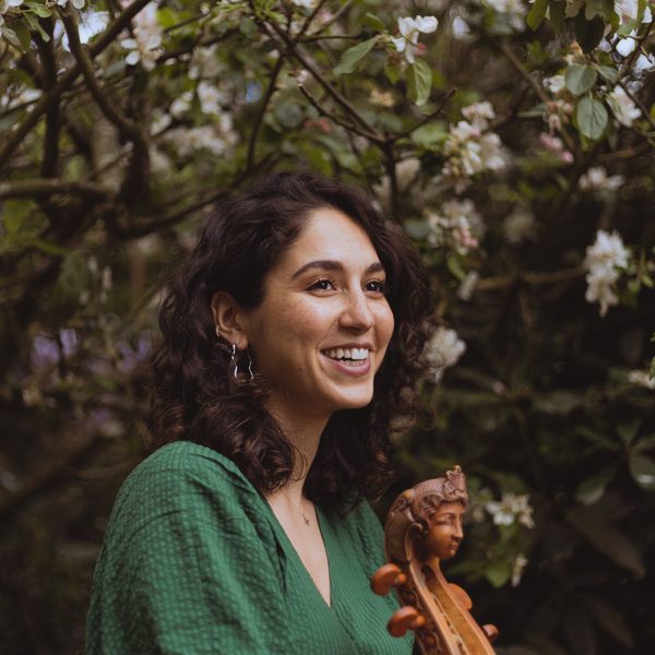 A woman in a green dress, pictured smiling, with the neck of a stringed instrument in the foreground and a tree in the background.