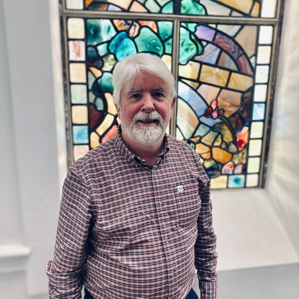 Luke Dunlea smiles, posing for camera in front of a stained glass window indoors