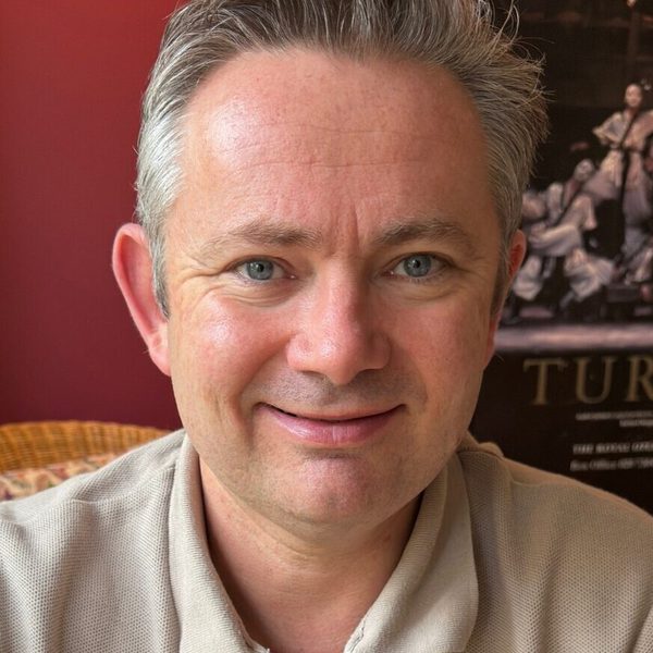 Matthew Dickinson smiles to camera backed by a dark red wall.