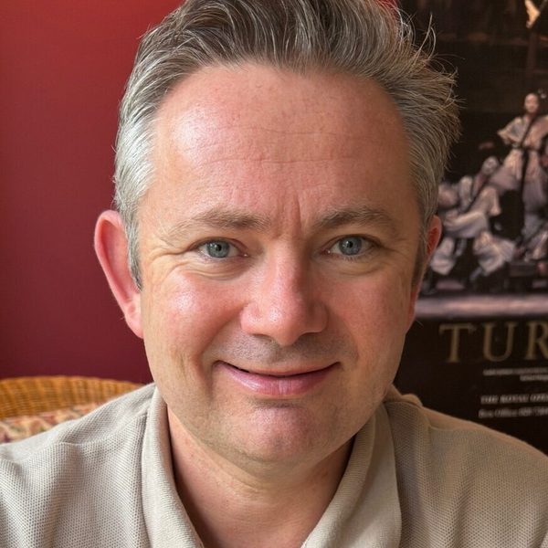 Matthew Dickinson smiles to camera backed by a dark red wall.