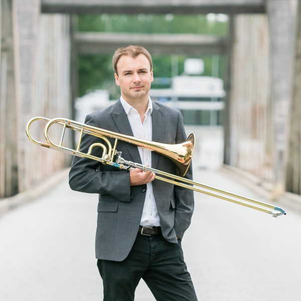 Matthew Gee poses for camera in an urban environment, holding a trombone.