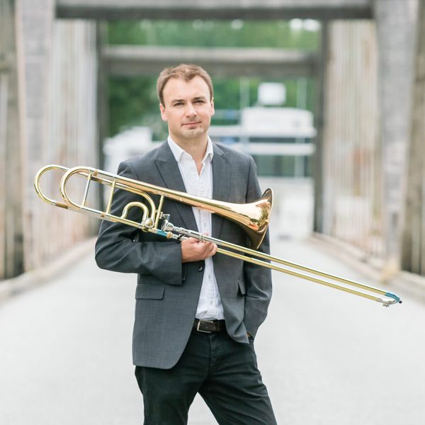 Matthew Gee poses for camera in an urban environment, holding a trombone.