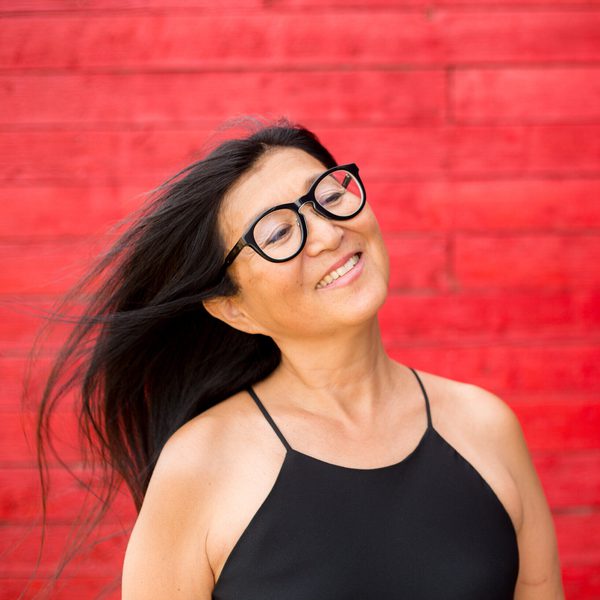 A smiling Megumi Masaki in profile, head tilted to the right, with a bright red-brick backdrop.
