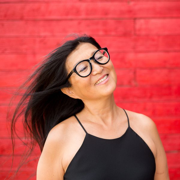 A smiling Megumi Masaki in profile, head tilted to the right, with a bright red-brick backdrop.