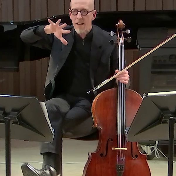 A man seated with a cello and bow supported in one hand, gesturing to speak with the other hand.