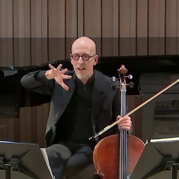 A man seated with a cello and bow supported in one hand, gesturing to speak. with the other hand.