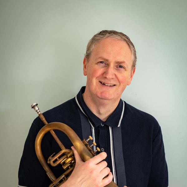 A smiling Nick Smart holds a trumpet, wearing a navy blue t-shirt and standing against a grey backdrop.