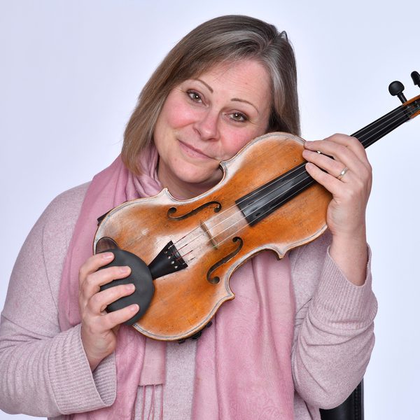 A woman in a pink sweater holds a violin up to her face, smiling to camera.