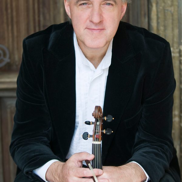 Pavlo Beznosiuk Headshotholds a violin at the neck with both hands, smiling to camera.