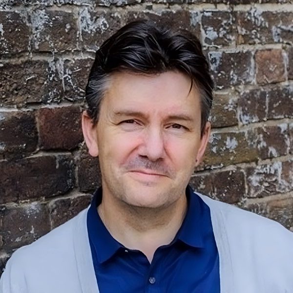Portrait of Pete Churchill smiling to camera, standing against a brick wall.