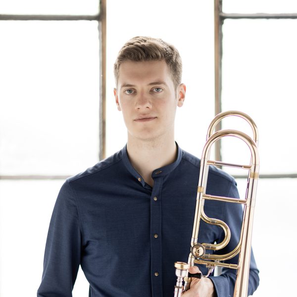Peter Moore poses with one hand in pocket and the other holding a trombone.