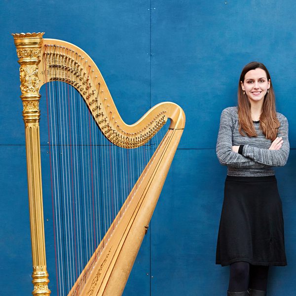 Rachel Wick stands against a blue background next to a harp, crossing arms.