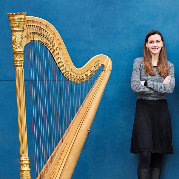 Rachel Wick stands against a blue background next to a harp, crossing arms.