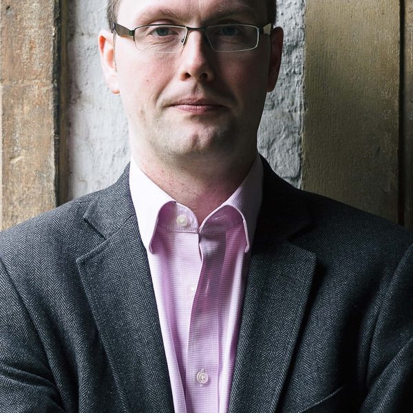 Portrait of Richard Pinel looking to camera, standing against a brick wall.