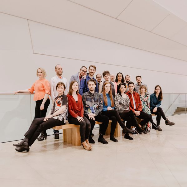 The Riot Ensemble sit across a bench, with the remainder of the group in a row behind, smiling to Camera without instruments.