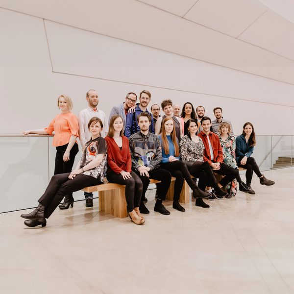 The Riot Ensemble sit across a bench, with the remainder of the group in a row behind, smiling to Camera without instruments.