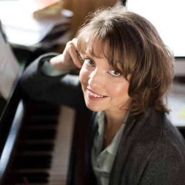 Roxanna Panufnik sits at a piano, smiling to camera.
