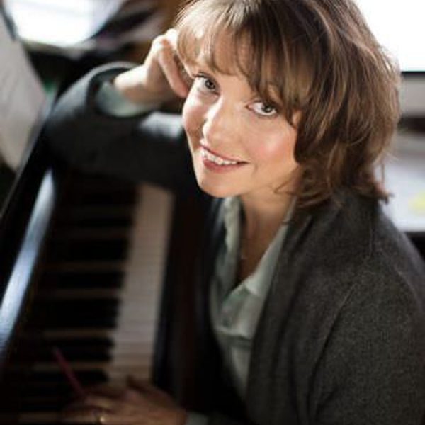 Roxanna Panufnik sits at a piano, smiling to camera.