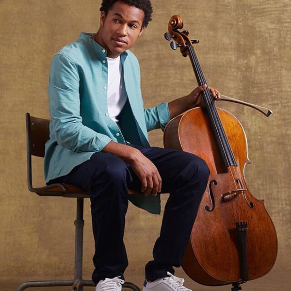 Sheku Kanneh-Mason sits holding his cello, facing camera.
