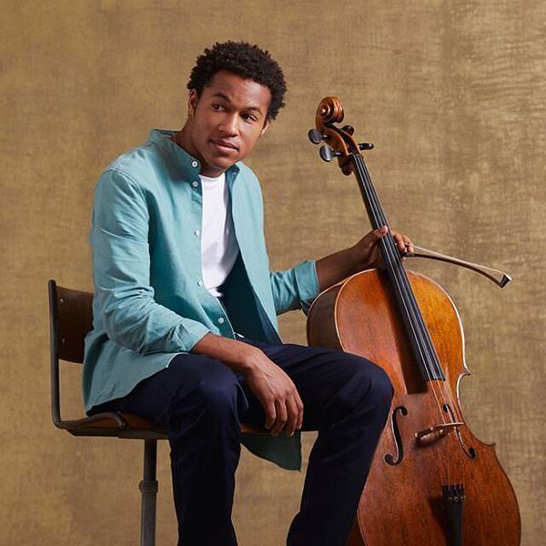 Sheku Kanneh-Mason sits holding his cello, facing camera.