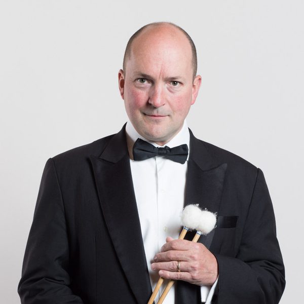 Simon Carrington wears a dinner jacket and looks to camera while holding drum beaters