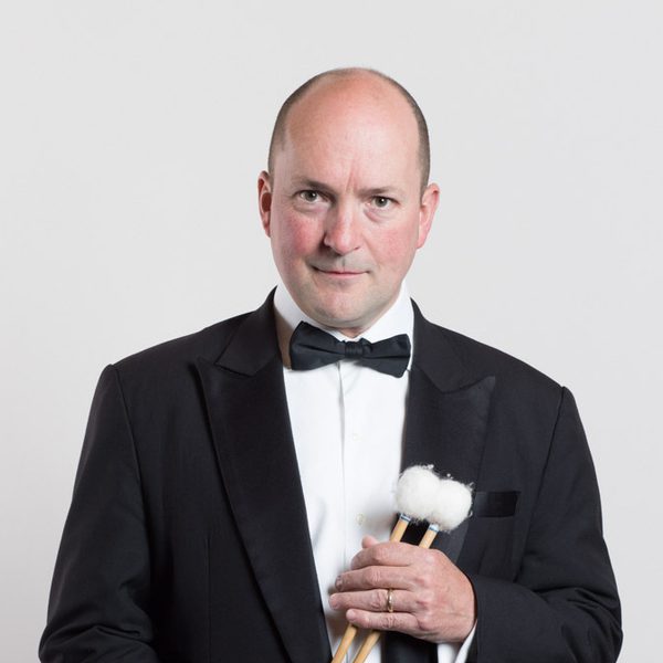 Simon Carrington wears a dinner jacket and looks to camera while holding drum beaters.