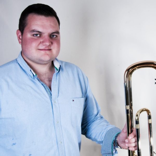 Simon Minshall holds a trombone posing for camera against a white wall.
