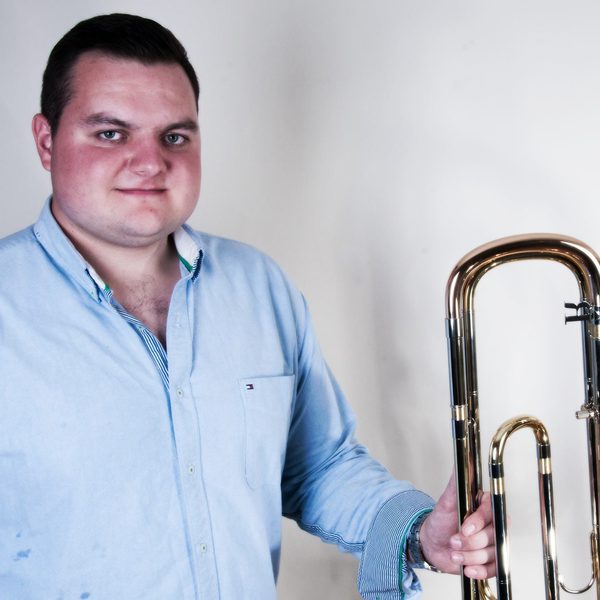 Simon Minshall holds a trombone posing for camera against a white wall.
