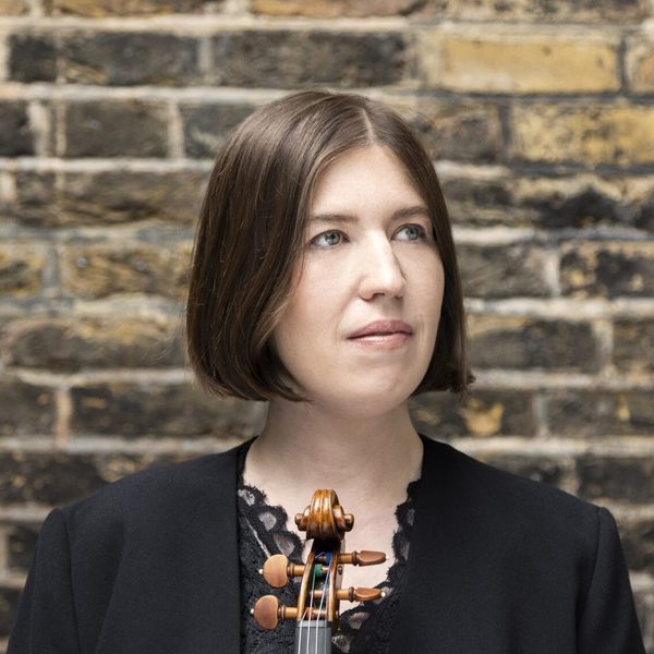 Portrait of Sini Simonen standing against a brick wall holding a violin, looking out of frame.