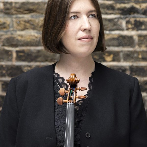 Portrait of Sini Simonen standing against a brick wall holding a violin, looking out of frame.