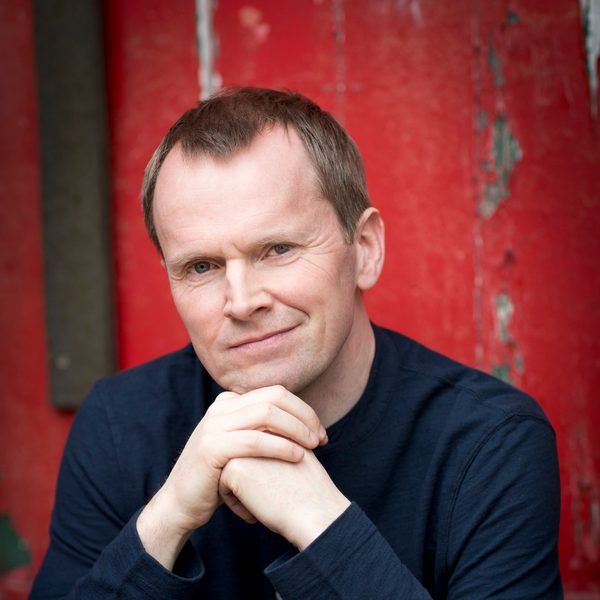 Steven Osborne sits outside in front of a red wall, posing for camera.