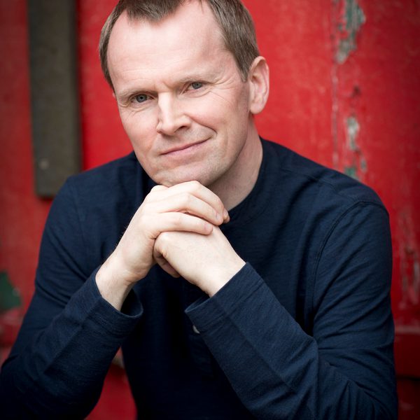 Steven Osborne sits outside in front of a red wall, posing for camera.