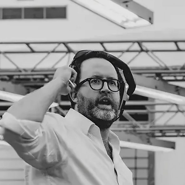 Tom Kelly stands on stage, lifting headphones from ear mid-discussion.