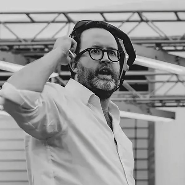 Tom Kelly stands on stage, lifting headphones from ear mid-discussion.