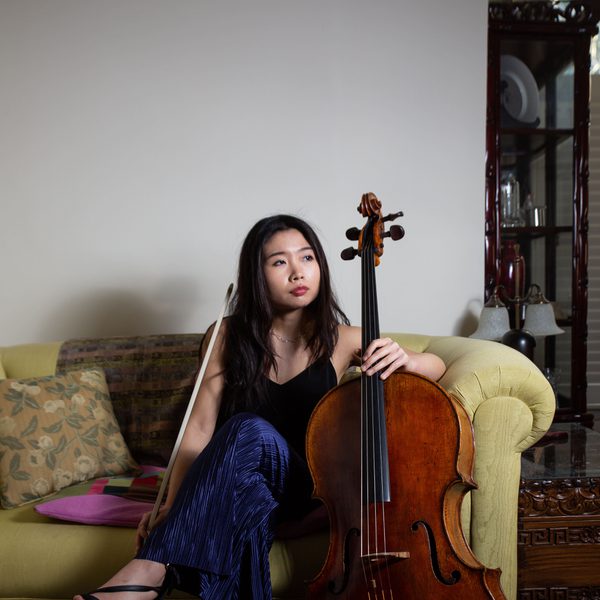Yurie Lee sits on a sofa holding cello upright, looking out of a window.