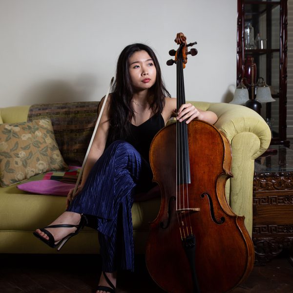 Yurie Lee sits on a sofa holding cello upright, looking out of a window.