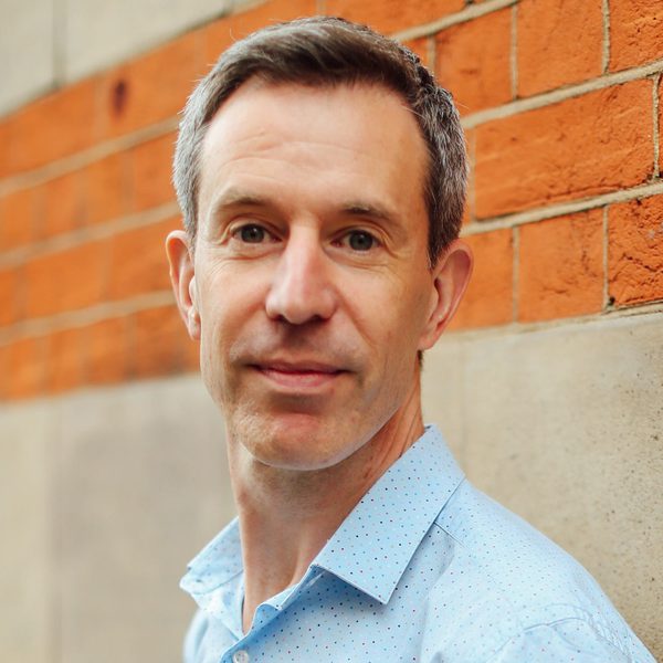 Portrait of Julian West looking to camera, smiling, against a brick wall