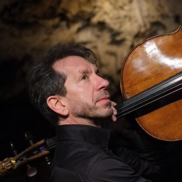 Robert Cohen holds a cello aloft over his shoulder, looking upwards.